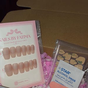May include: A set of nude-colored press-on nails in a clear plastic sleeve with a pink card labeled NAILS BY FATIMA, alongside a clear bag containing adhesive tabs, a Star alcohol swab, and a small orange nail file inside a cardboard box.