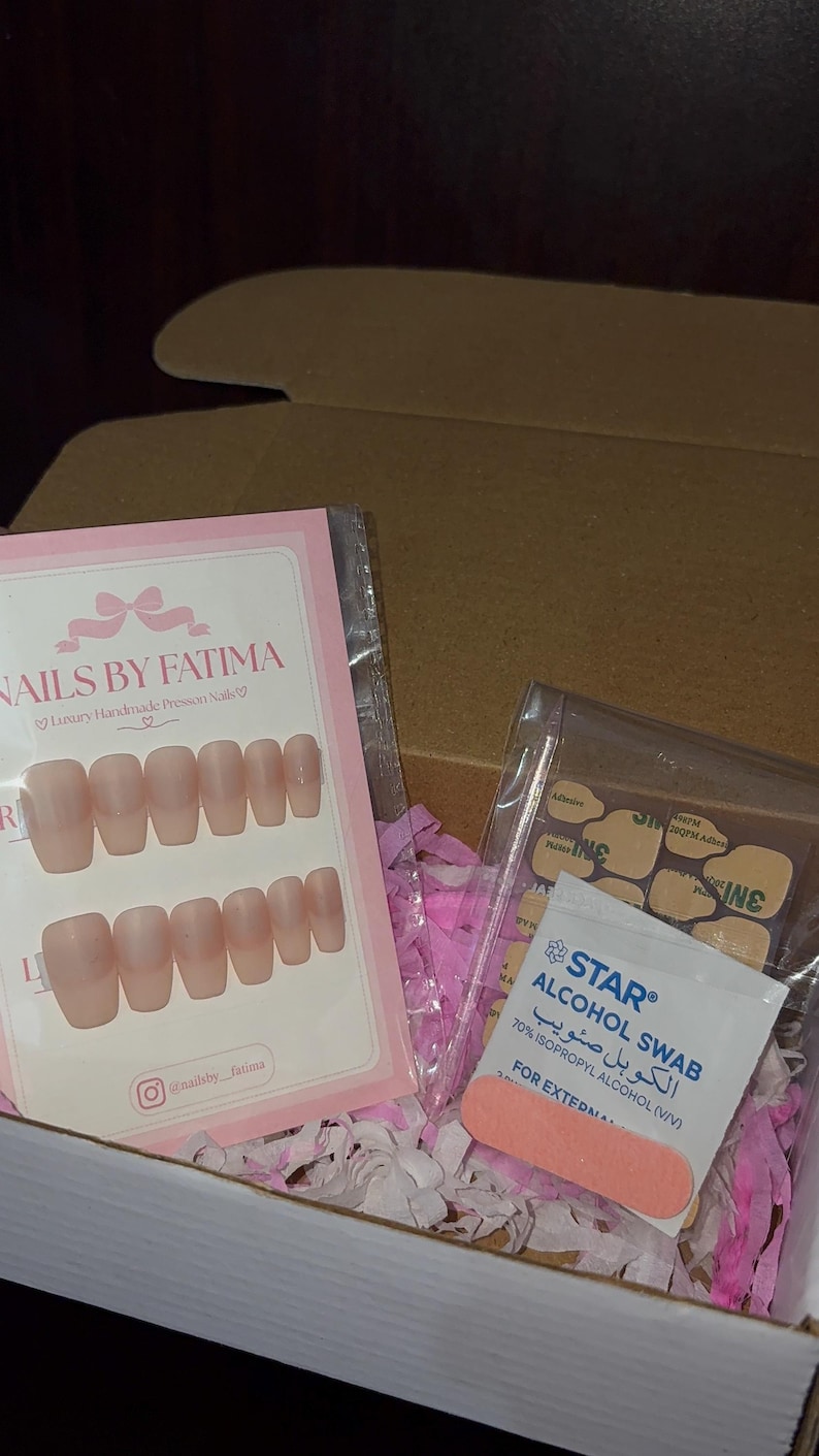 May include: A set of nude-colored press-on nails in a clear plastic sleeve with a pink card labeled NAILS BY FATIMA, alongside a clear bag containing adhesive tabs, a Star alcohol swab, and a small orange nail file inside a cardboard box.