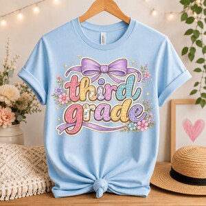 Op de afbeelding: Een lichtblauw t-shirt met korte mouwen en een grafische print met de tekst "third grade" in kleurrijke, gedessineerde bubbelletters. Het ontwerp bevat een grote paarse strik aan de bovenkant en kleine bloemaccenten rondom de tekst.