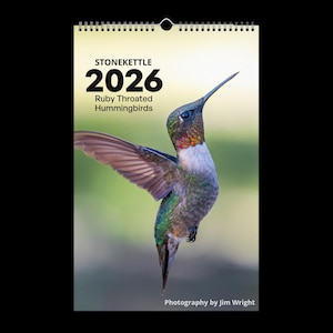 May include: A calendar for 2026 featuring a photograph of a Ruby-Throated Hummingbird in flight. The calendar has a spiral binding at the top and includes the text "STONEKETTLE 2026 Ruby Throated Hummingbirds" and "Photography by Jim Wright". The hummingbird has iridescent green, red, and white feathers.