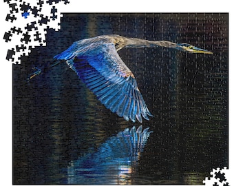 May include: A completed jigsaw puzzle featuring a great blue heron in flight over water. The bird's blue and gray feathers are detailed, and its reflection is visible in the water. Some puzzle pieces are separate.