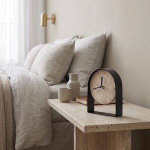 May include: A table clock featuring a circular beige travertine stone face with black hands, encased within a matte black metal arch frame. The clock has a clean, structural design with no numerals on the face, highlighting the natural texture of the stone.