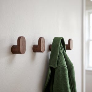 May include: Three J-shaped wall hooks made of dark brown wood with a visible grain pattern. The hooks are mounted horizontally in a row on a plain white wall. A green wool coat hangs from the third hook on the right side of the wall arrangement.