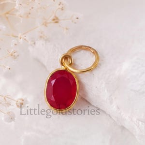 Puede incluir: Un colgante de oro con una piedra preciosa de color rojo intenso, forma ovalada y tallada, engastada en un bisel de oro liso con una anilla circular en la parte superior. El texto Littlegoldstories aparece impreso en letras negras debajo.