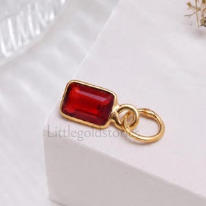 Puede incluir: Un colgante de tono dorado con una piedra preciosa rectangular de color rojo intenso engastada en un marco de metal liso con una anilla circular. El texto Littlegoldstones aparece grabado sutilmente en la superficie junto a la joya.