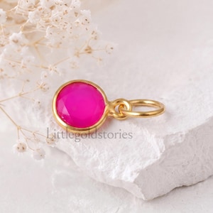 Könnte beinhalten: Ein runder, facettierter, leuchtend pinker Edelsteinanhänger in einer goldfarbenen Metallfassung mit einem befestigten Biegering. Der Text Littlegoldstories ist auf der Oberfläche zu sehen. Das Schmuckstück liegt auf einem weißen, strukturierten Untergrund.
