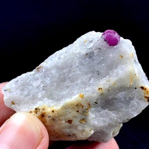 May include: A raw mineral specimen featuring a translucent, crystalline white rock matrix with a single, small, vibrant magenta-pink gemstone embedded near the top edge, displaying natural textures and irregular, jagged surface formations.