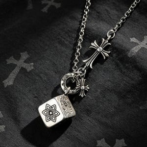 Pode incluir: Um colar de tom prateado com uma corrente e um conjunto de pingentes pendentes. O conjunto inclui um berloque em forma de cubo com uma gravação floral, um anel circular com símbolos gravados e dois berloques em forma de cruz.