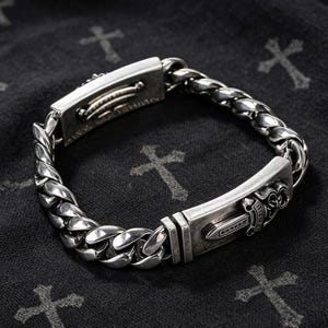 May include: A silver chain bracelet featuring a thick, polished curb link design. The bracelet includes two rectangular metal plates, one of which is engraved with a detailed dagger motif and ornate scrollwork, while the other shows text.