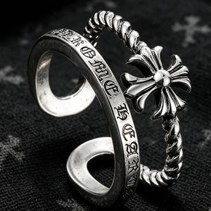May include: A silver-toned open ring featuring two distinct bands. One band is smooth with engraved black lettering, while the other is a twisted rope design topped with a polished, four-pointed cross emblem with flared ends.