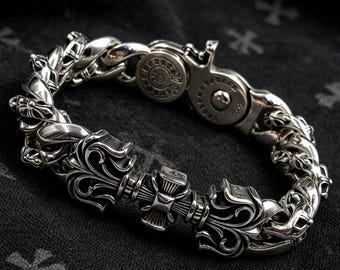 Puede incluir: Una pulsera de eslabones de plata de ley pulida con intrincados grabados en forma de flores y cruces. La pulsera incluye un mecanismo de cierre circular con texto grabado y una bisagra de seguridad, mostrando metal detallado.