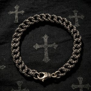Könnte beinhalten: Ein silbernes Kettenarmband mit sich wiederholenden, verzierten kreuzförmigen Gliedern. Das Armband wird mit einem Karabinerverschluss gesichert, der kleine Gravurdetails aufweist. Das Metall hat ein poliertes Finish mit dunklen Vertiefungen.