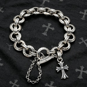 Puede incluir: Una pulsera de eslabones de plata con anillos circulares grabados con texto, un cierre circular central con el texto 2003 Sterling, un dije de cruz colgante y una pequeña cadena adicional sujeta al mecanismo del cierre.