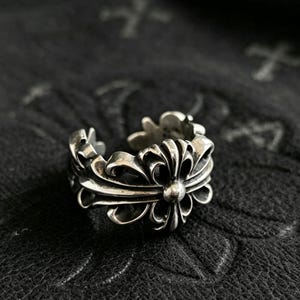 May include: A silver-toned metal ring featuring an open band design with a prominent floral-inspired motif. The surface of the ring displays intricate, symmetrical scrollwork and curved detailing with darkened recessed areas to highlight the metal texture.