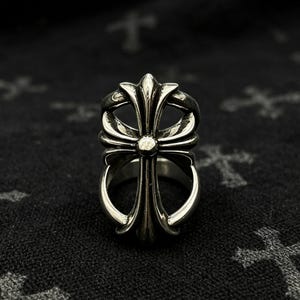 Puede incluir: Un anillo de metal plateado con un diseño calado centrado en un motivo de cruz prominente. La cruz tiene brazos redondeados y ensanchados con un acabado pulido y liso que contrasta con las áreas oscuras y hundidas de la banda metálica.