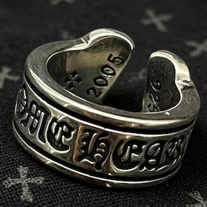 May include: A silver open-band ring featuring black engraved gothic-style lettering around the exterior. The interior of the band is stamped with the year 2005, a small cross symbol, and the number 925, indicating sterling silver composition.