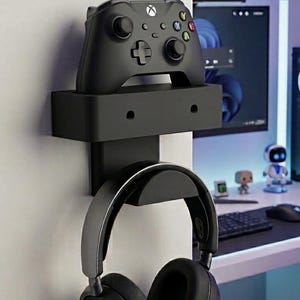 Könnte beinhalten: Eine schwarze Wandhalterung, die oben einen schwarzen Gamecontroller und darunter einen schwarzen Over-Ear-Kopfhörer hält. Der Text MODERN BRACKET DESIGN und SECURE. SLEEK. ESSENTIAL. ist sichtbar. Die Halterung hat zwei kreisförmige Schraublöcher.