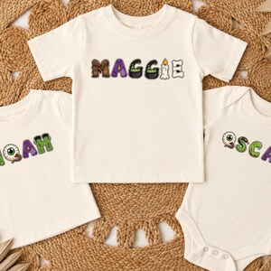 以下が含まれることがあります： オフホワイトの衣類3点。半袖Tシャツ2枚とベビー用ボディスーツ1枚です。各シャツには、目玉、大釜、骨などのハロウィーンをテーマにしたカラフルな文字グラフィックで、名前がデザインされています。