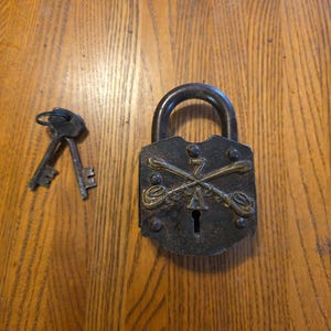 Puede incluir: Un candado antiguo de metal oscuro con un ojo de cerradura y un asa curva. La cerradura presenta espadas cruzadas decorativas y una llave se coloca junto a ella sobre una superficie de madera. El candado es probablemente vintage.