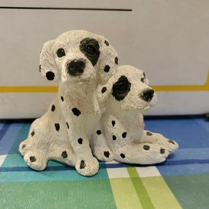 以下が含まれることがあります： 2つの小さなセラミック製のダルメシアン犬の置物。1つは後ろに座っています。犬は白地に黒の斑点があり、黒い鼻と目を持っています。
