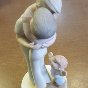May include: A porcelain figurine of a family with three figures. The two larger figures are a woman and a child, both with blonde hair, hugging each other. The smaller figure is a child with brown hair standing in front of the other two. The base of the figurine is white and has the words "My Turn Next" written on it.