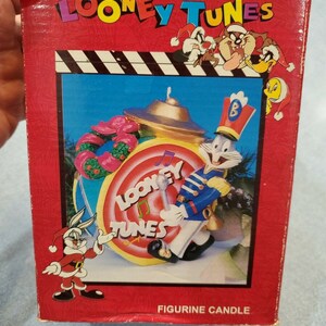 May include: A red box with the words "Looney Tunes" printed in white. The box features a cartoon image of Bugs Bunny dressed as a drummer, playing a drum with the words "Looney Tunes" on it. The box also features other Looney Tunes characters, including Daffy Duck, Tweety Bird, and Sylvester the Cat. The box says "Figurine Candle" on the bottom.
