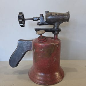 Op de afbeelding: Een antieke rode en zwarte brander. De brander heeft een rode cilindrische brandstoftank, een zwarte handgreep en een zwarte bedieningsknop. Het bovenste gedeelte is zwart met een mondstuk. De brander is waarschijnlijk van metaal.