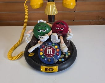 Könnte beinhalten: Ein neuartiges Telefon mit gelbem Hörer und Sockel, verbunden durch ein Spiralkabel. Der schwarze Sockel ist mit grünen und roten M&M's-Figuren verziert. Das Ziffernfeld hat bunte Tasten und die Aufschrift "REDIAL M FLASH". Das Telefon hat das "m-m"-Logo.