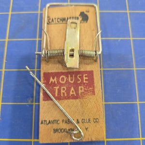 Könnte beinhalten: Eine Mausefalle aus Holz mit einem Metallfedermechanismus und einem Drahtbügel. Die Holzbasis trägt die Aufschrift CATCHMASTER, MOUSE TRAP und ATLANTIC PASTE & GLUE CO. BROOKLYN, N.Y. Ein Metallauslöser erstreckt sich diagonal über den unteren Teil.