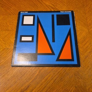 May include: A vinyl record album cover with a geometric design. The cover features a blue background with orange triangles, black rectangles, and white squares. The text "SPLIT ENZ" and "TRUE COLOURS" are visible.
