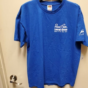May include: A royal blue t-shirt with a white graphic that says "Rascal Flatts Local Crew Here's To You Tour". The shirt has a small logo on the right sleeve.