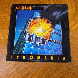 Könnte beinhalten: Das Albumcover von Def Leppard's "Pyromania". Das Artwork zeigt ein Fadenkreuz, das über einer feurigen Explosion liegt, vor dem Hintergrund eines modernen Gebäudes. Der Bandname steht in gelber, eckiger Schrift oben. Der Albumtitel ist unten gelb.