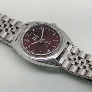 May include: A silver-tone wristwatch with a dark maroon dial featuring white Devanagari numerals and silver hands. The watch has a polished stainless steel case and a multi-link metal bracelet. The dial includes the text HMT and Kohinoor.