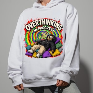 May include: A white hooded sweatshirt featuring a graphic of a sloth wearing a black hoodie and tan pants, lying on a pile of colorful pillows. Above the sloth, the text reads OVERTHINKING IN PROGRESS against a vibrant rainbow spiral background.