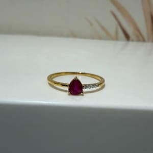 May include: A gold ring featuring a pear-shaped red ruby gemstone as the central focal point. The band includes a small section of three tiny, clear, round-cut accent stones set into the gold shank adjacent to the ruby setting.