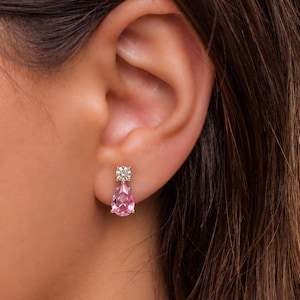 Peut inclure: Une boucle d'oreille pendante composée d'une petite pierre ronde transparente à facettes en haut, reliée à une pierre plus grande en forme de poire, rose clair et à facettes en bas. Les pierres sont serties dans une monture métallique.