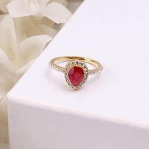Peut inclure: Une bague en or ornée d'une pierre rouge en forme de poire au centre, entourée d'un halo de petites pierres claires. L'anneau est également agrémenté de petites pierres claires sur les côtés, offrant un aspect brillant et réfléchissant.