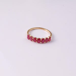 May include: A gold ring featuring a row of six oval-cut, deep red gemstones set in a horizontal line. The band is thin and polished, showcasing a simple metal construction that holds the vibrant red stones securely in a prong setting arrangement.