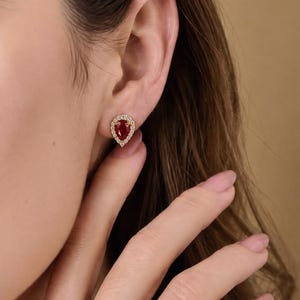 May include: A pear-shaped red gemstone stud earring set in a gold-toned metal frame, surrounded by a halo of small, clear, round-cut white stones. The earring is secured to the earlobe with a post backing, showcasing a classic teardrop design.