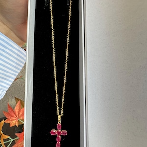 Peut inclure: Un collier à chaîne dorée avec un pendentif en forme de croix. Le pendentif est composé de cinq pierres précieuses ovales d'un rouge rosé profond disposées en croix, avec une petite bélière dorée reliant le pendentif à la fine chaîne métallique.