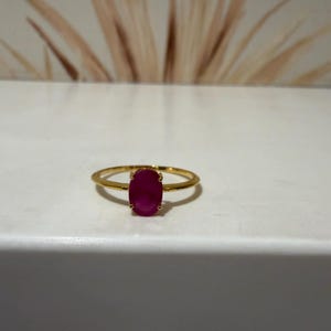 Puede incluir: Un anillo de oro con una piedra preciosa de color rosa intenso en forma ovalada, montada en un engaste de cuatro garras. El aro es fino y pulido, con un acabado metálico liso que complementa el color vibrante de la piedra central.