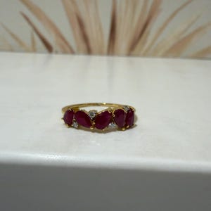 Puede incluir: Un anillo de oro con una hilera de cinco rubíes de color rojo intenso en forma de pera, alternados con pequeñas piedras de acento redondas y transparentes. La banda es fina y de oro pulido, mostrando una disposición simétrica de las gemas.