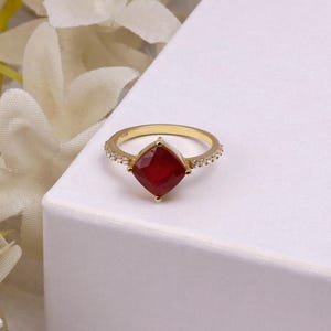 Peut inclure: Une bague en or avec une pierre précieuse rouge foncé taille coussin fixée par quatre griffes. L'anneau est orné de petites pierres claires taille ronde sur les côtés, créant un effet scintillant sur la finition en métal doré poli.