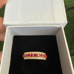 Könnte beinhalten: Ein goldener Bandring mit einer mittleren Reihe aus runden roten Rubinen, flankiert von zwei Reihen kleiner, klarer weißer Diamanten. Der Ring wird in einer weißen Schmuckschatulle mit schwarzem Samtfutter präsentiert, die den Besatz zeigt.