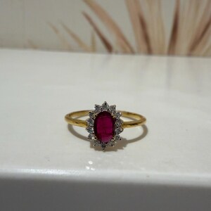 May include: A gold ring featuring a central oval-shaped deep red gemstone surrounded by a halo of small, clear, sparkling stones. The band is thin and polished, providing a classic look that highlights the vibrant contrast between the red centre and the surrounding stones.