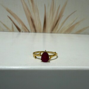 Pode incluir: Um anel de ouro com uma pedra preciosa vermelho-escura em forma de pera no centro. A banda do anel tem um design distinto que lembra ramos finos ou chifres que se estendem a partir da base da cravação central da pedra.