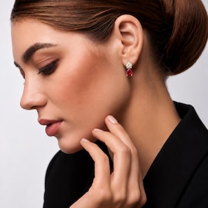 May include: A gold-toned earring featuring a cluster of small, clear, round-cut stones at the top, from which a single, teardrop-shaped, deep red gemstone hangs. The earring is secured to the earlobe with a post-style backing.