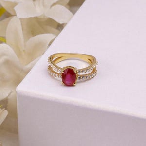 Peut inclure: Une bague en or avec une pierre précieuse rouge centrale de taille ovale maintenue par quatre griffes. L'anneau est divisé en deux rangées parallèles, chacune ornée de petites pierres claires rondes qui s'étendent sur les côtés de la bague.
