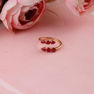 May include: A gold open-style ring featuring two clusters of three pear-shaped red gemstones. The ring has a smooth, polished gold band that curves around the finger, with each cluster of stones positioned at the open ends of the band.
