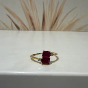 Op de afbeelding: Een gouden ring met twee rechthoekige, dieprode robijnen die verticaal in het midden zijn geplaatst. De ring is geaccentueerd met twee kleine, heldere, rondgeslepen diamanten aan weerszijden van de robijnen op een dunne gouden band.